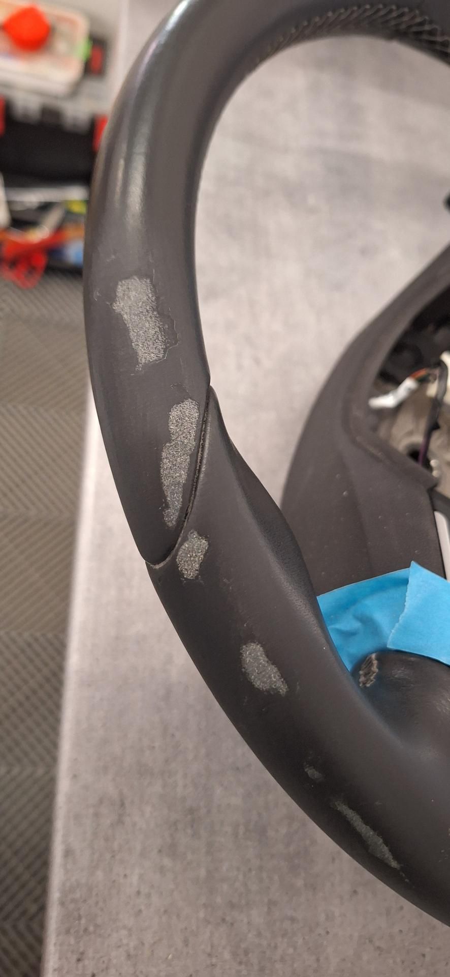 Gros plan sur un volant noir endommagé. Des zones d'usure sont visibles sur la poignée. Du ruban adhésif bleu est également présent.