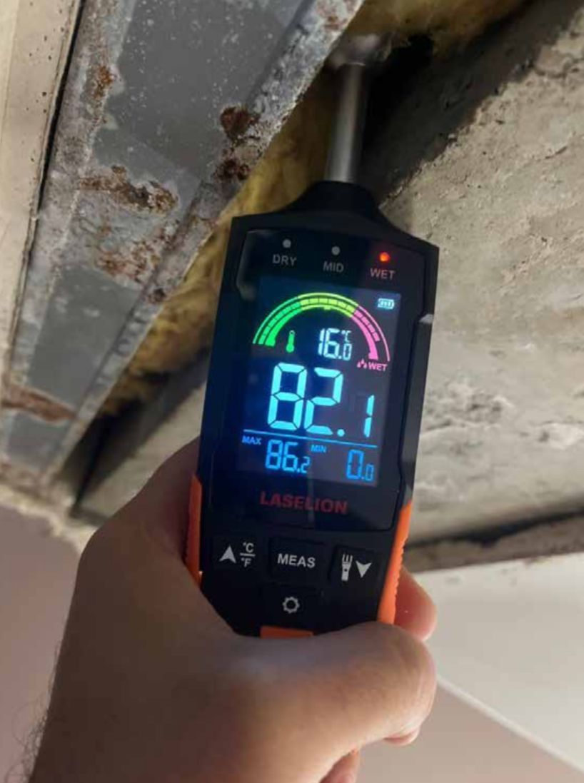 Une main tient un hygromètre contre le plafond. L'appareil affiche le taux d'humidité et la température.