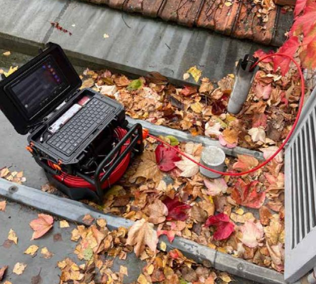 Un ordinateur portable et un câble rouge sont posés sur un toit recouvert de feuilles, à côté d'un tuyau en béton et de feuilles rouges.