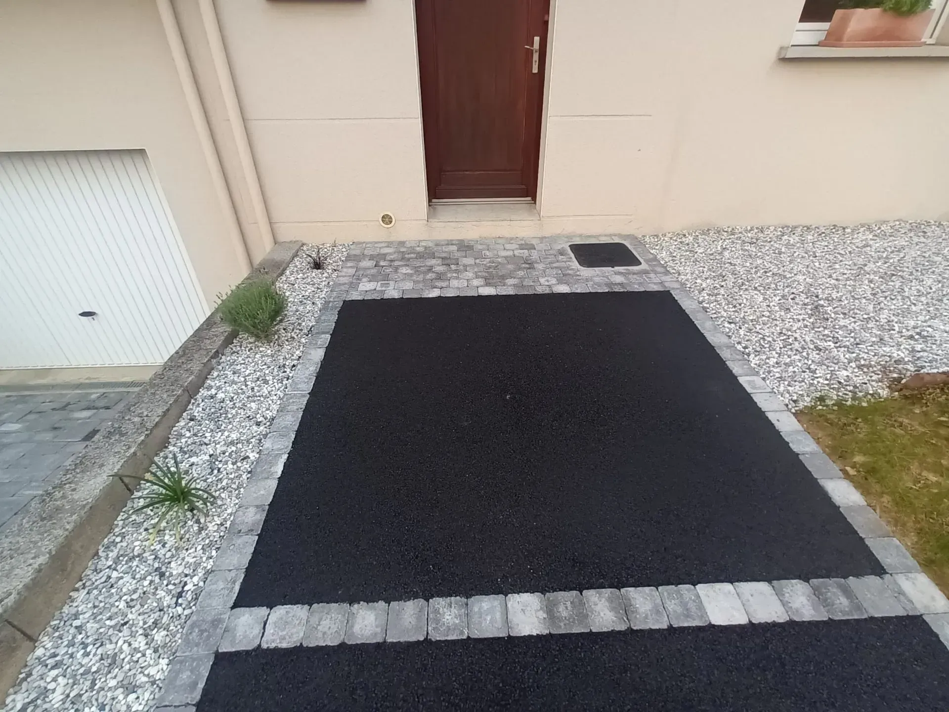 Une allée noire bordée de pavés gris et de gravier blanc mène à une porte marron.