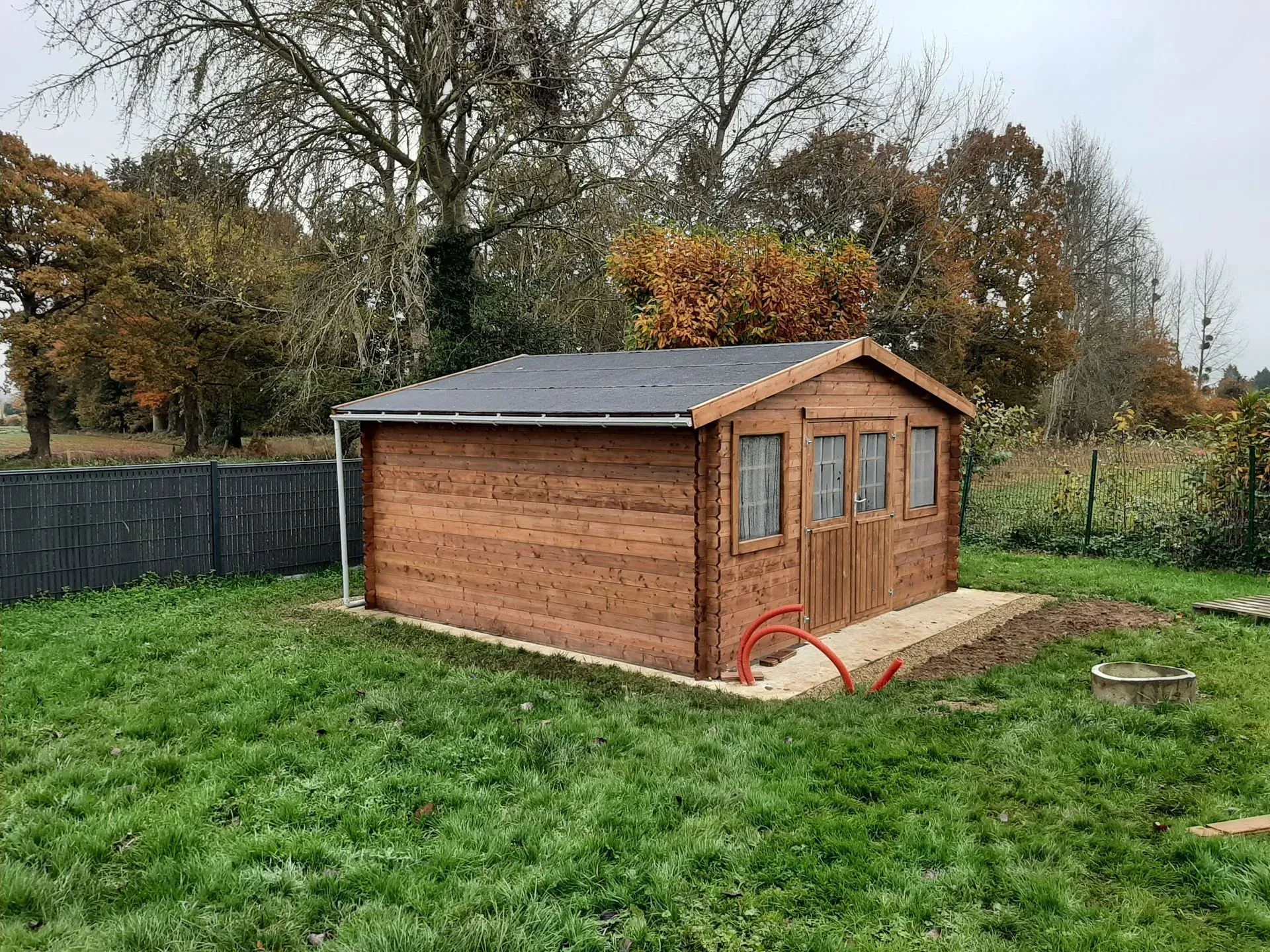 Abri en bois dans une cour herbeuse, marron avec un toit sombre. Arbres et clôture en arrière-plan.