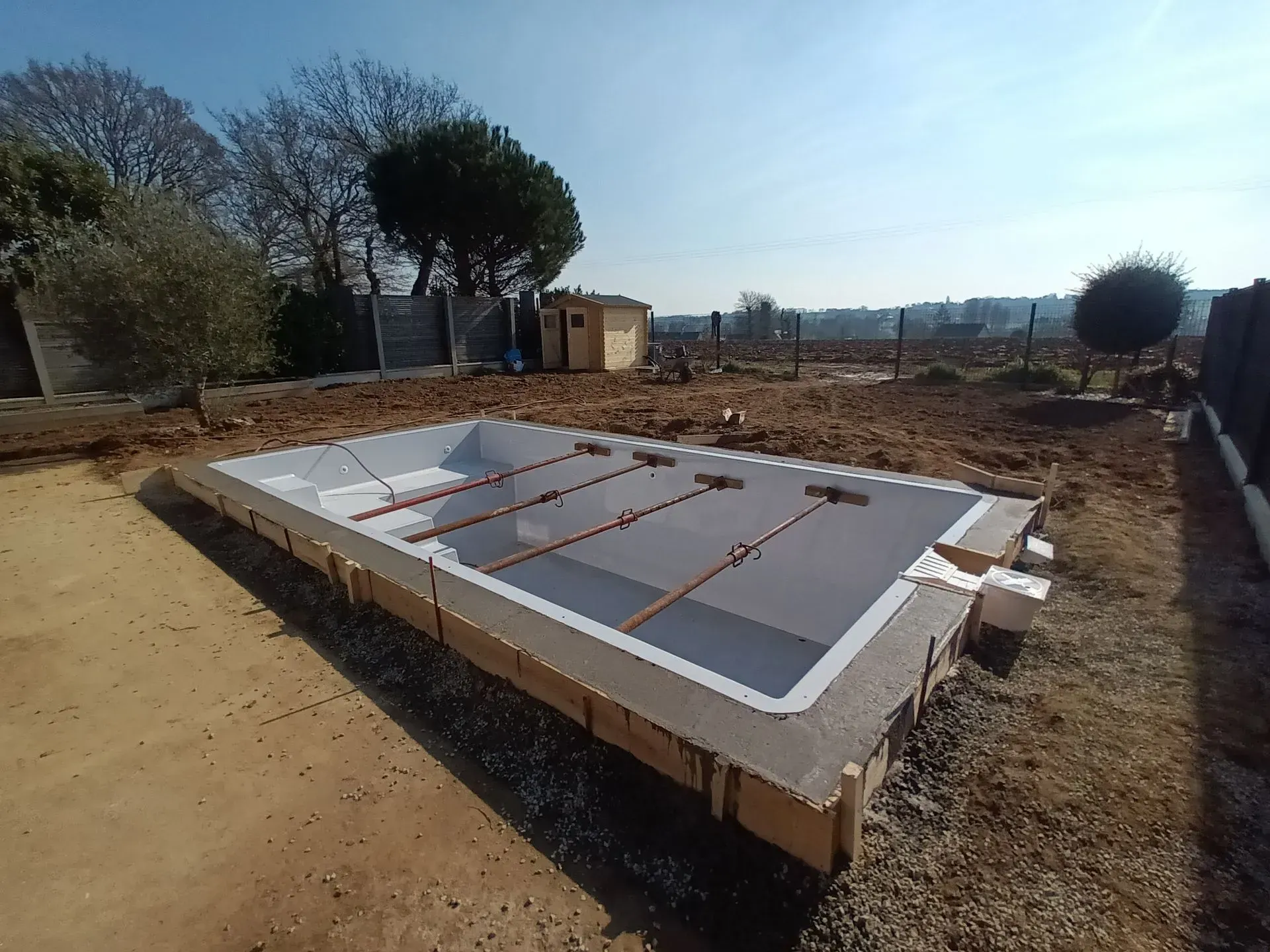 Une piscine rectangulaire en construction dans un jardin. Un sol brun entoure le bassin blanc et gris.