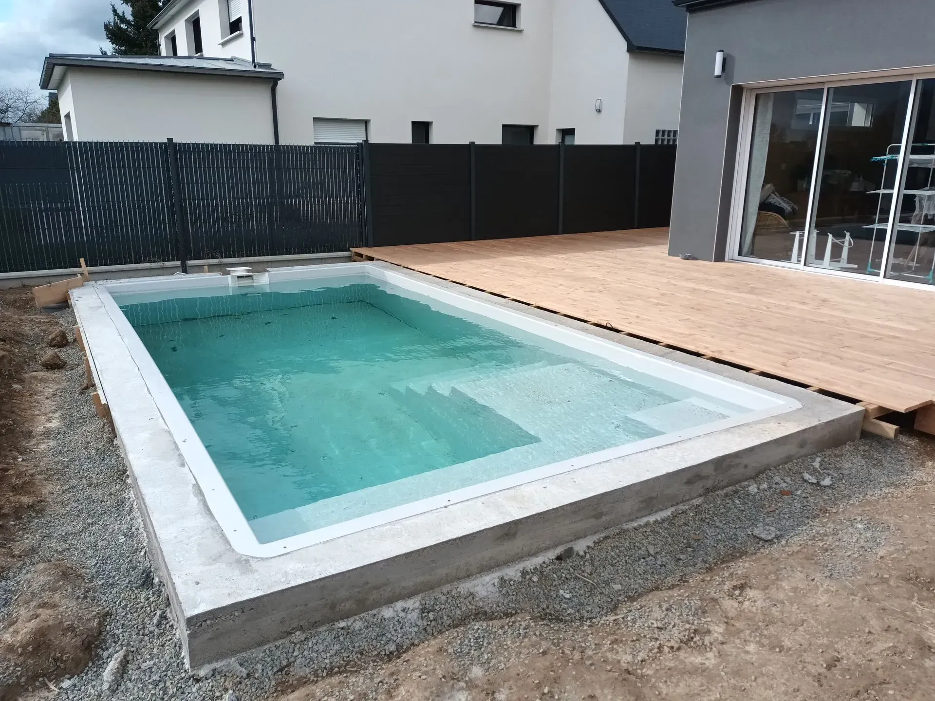 Piscine rectangulaire avec marches, entourée de béton et d'une terrasse en bois, dans un cadre arrière.