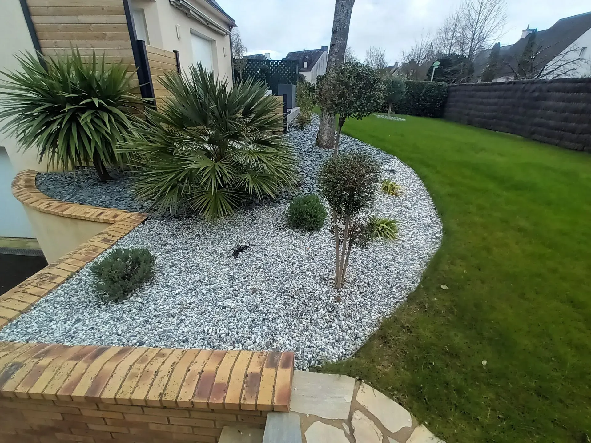 Cour avant paysagée avec gravier gris, plantes vertes et bordures en briques et pierres.