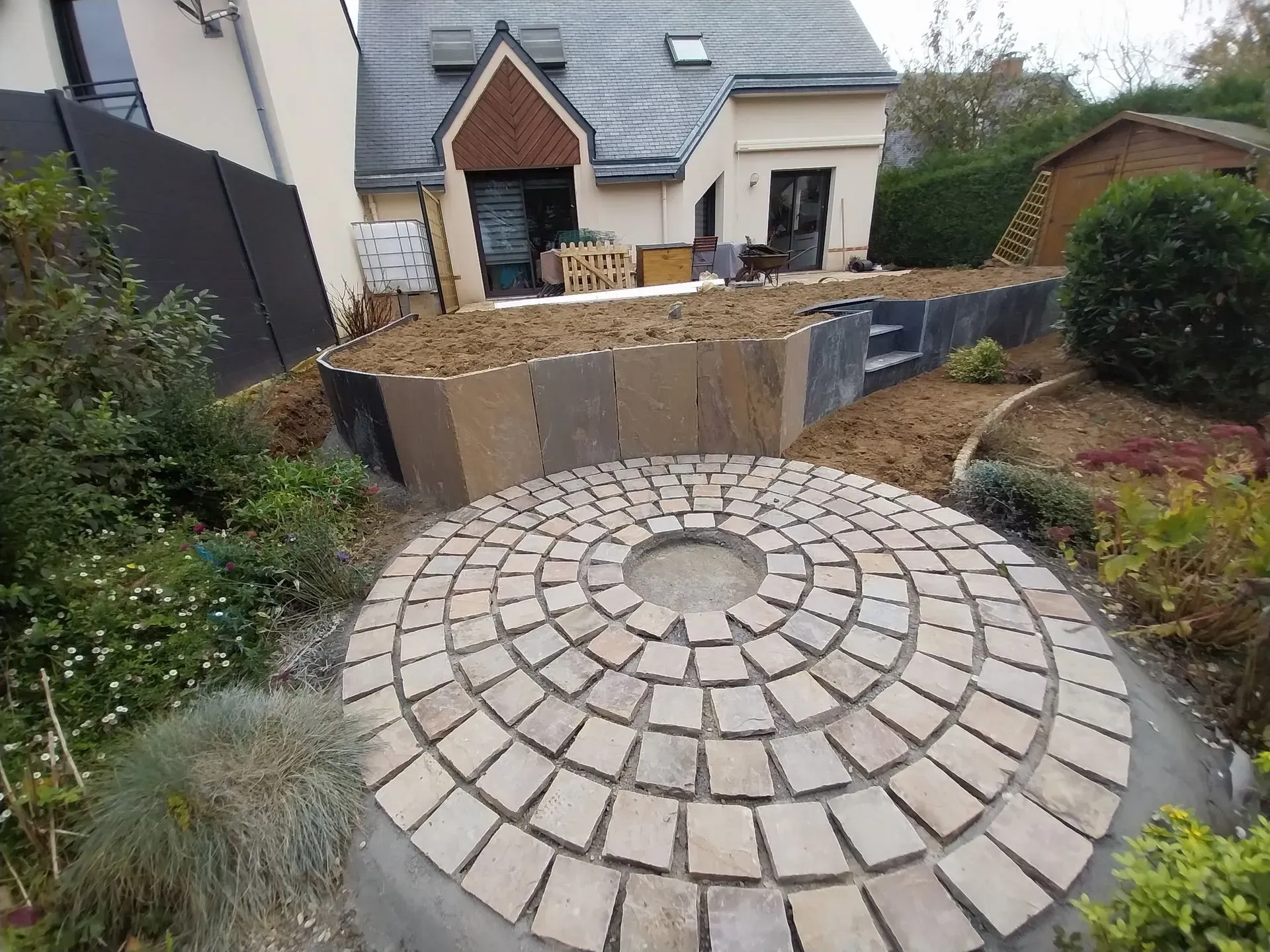 Patio circulaire en pierre avec un trou au milieu, adjacent à un jardin paysager et à une maison.