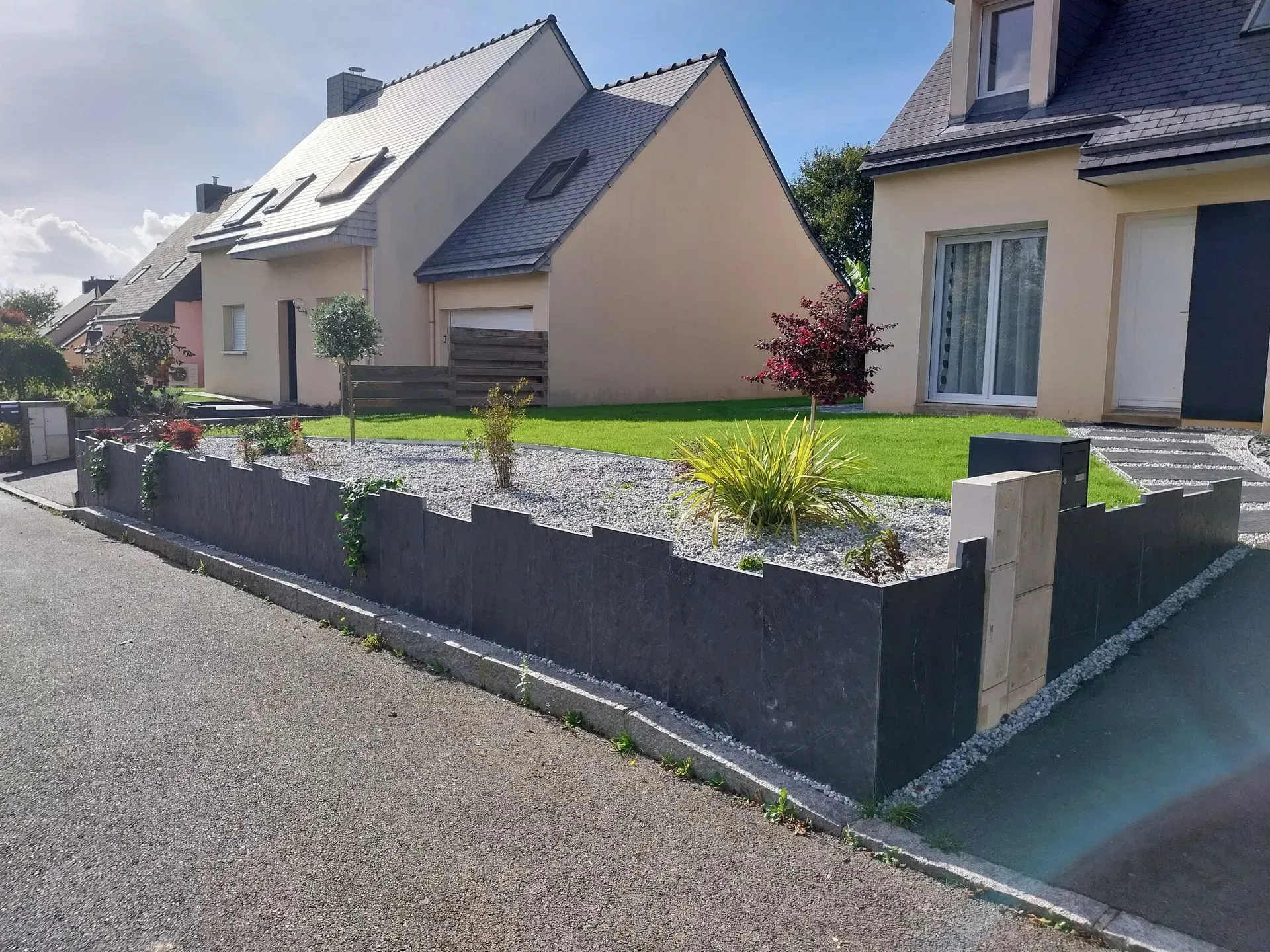 Muret gris bas avec des plantes et du gravier devant des maisons à deux étages dans une rue avec un ciel clair.