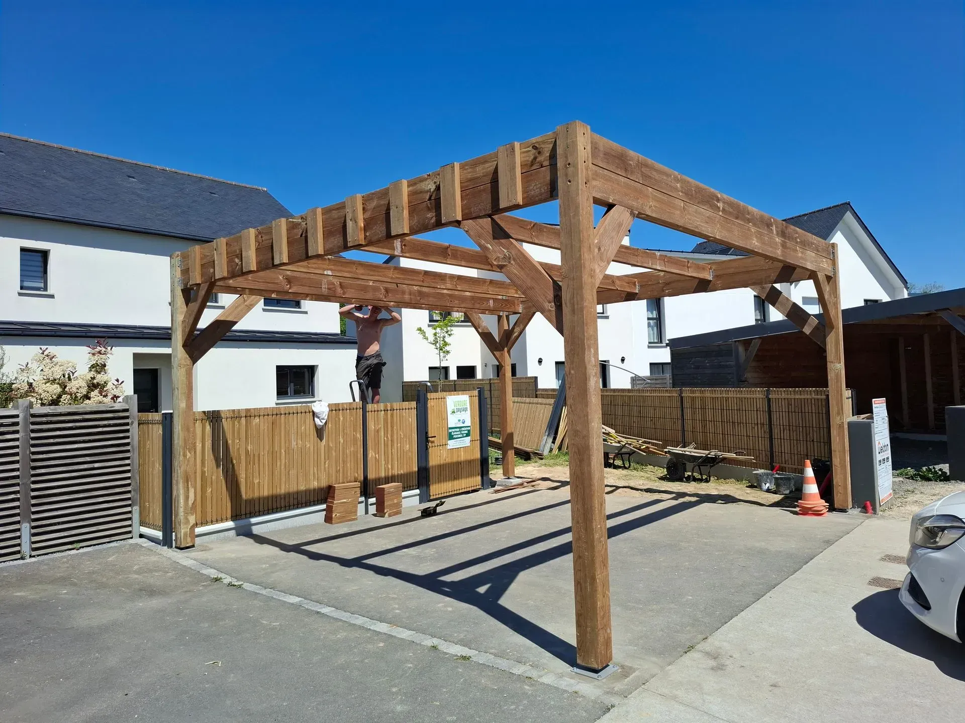 Abri de voiture en bois devant une maison, sous un ciel bleu clair. Une personne travaille à l'intérieur.