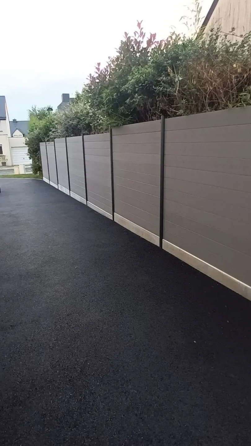 Clôture d'intimité grise le long d'une allée en asphalte foncé, avec des buissons verts au-dessus.
