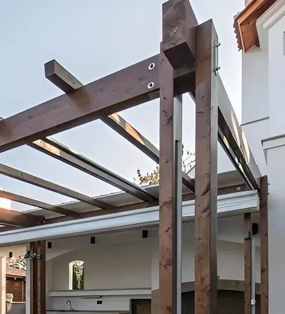Pergola en bois avec poutres teintées au-dessus d'un espace cuisine extérieur, attachée à un bâtiment blanc.