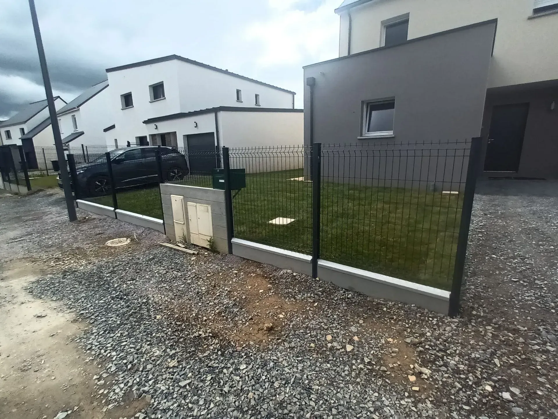 Une clôture noire entoure une petite cour devant une maison moderne grise et blanche avec un ciel nuageux.