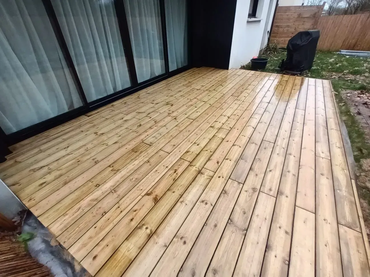 Terrasse en bois devant une maison, mouillée par la pluie. Grandes portes vitrées visibles.