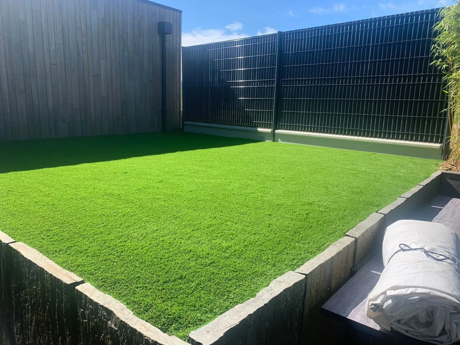 Gazon artificiel vert dans une cour arrière clôturée, clôture marron, mur gris, couverture enroulée, soleil éclatant.