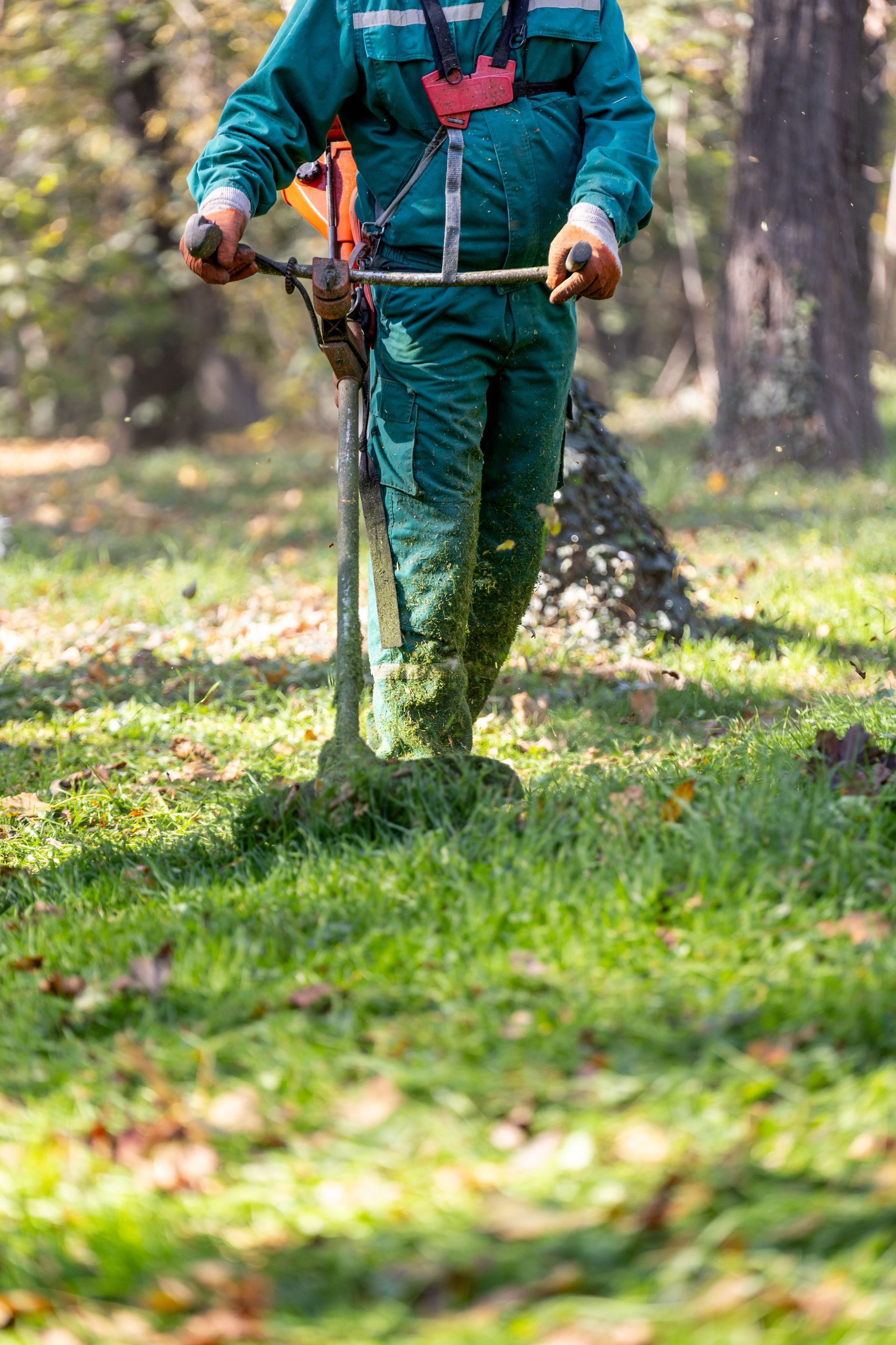 Une personne en vêtements de travail verts utilise un coupe-bordures pour tondre l'herbe dans un parc.