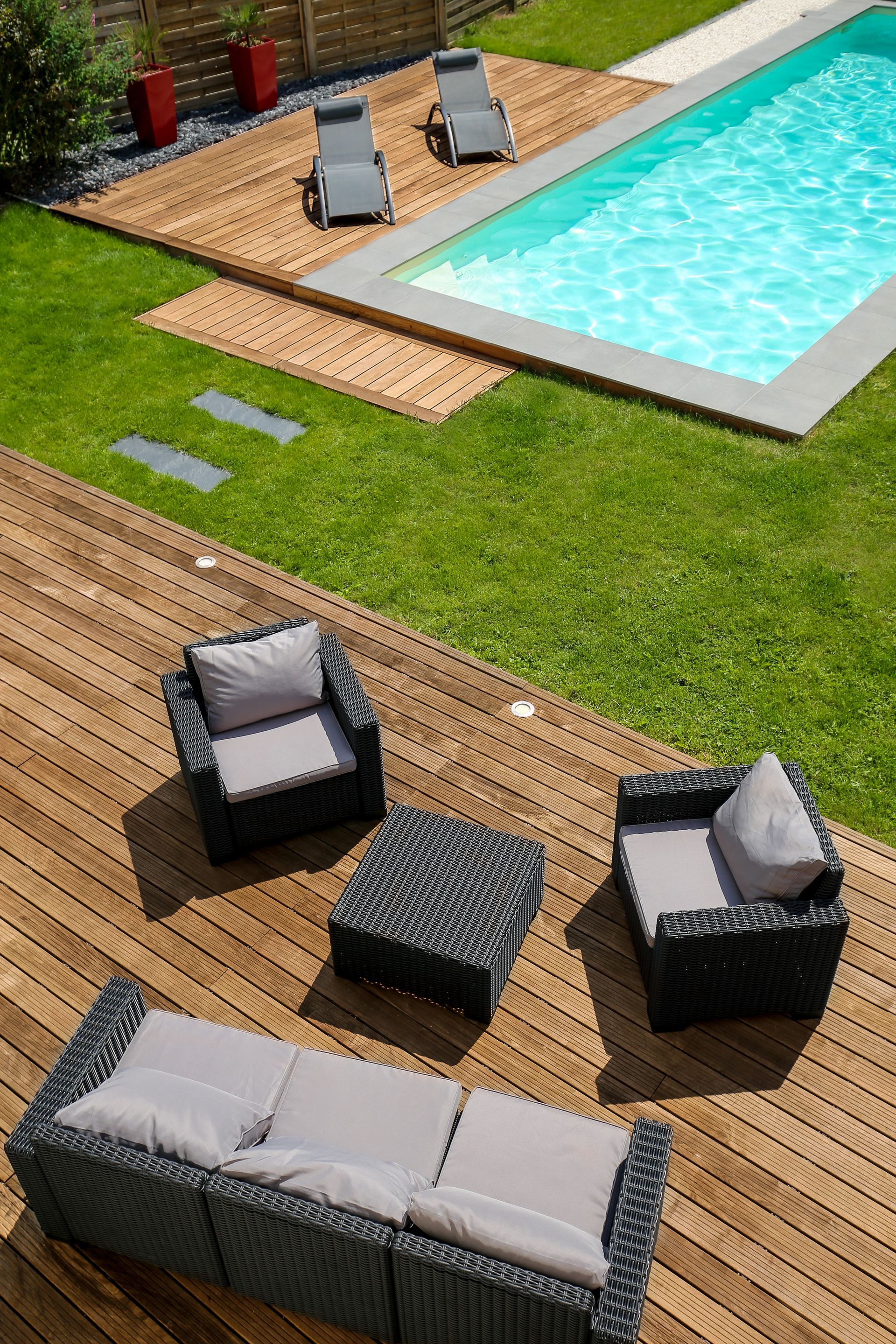 Terrasse en bois avec mobilier en osier, piscine et chaises longues lors d'une journée ensoleillée.