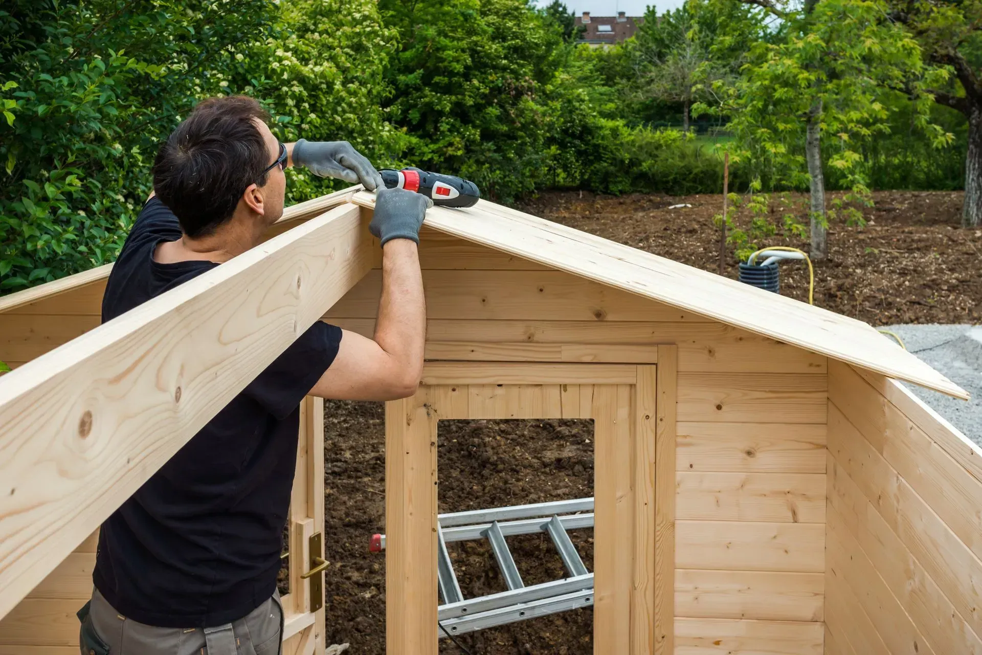 Un homme portant des gants construit un toit de hangar en bois avec une perceuse dans un jardin.