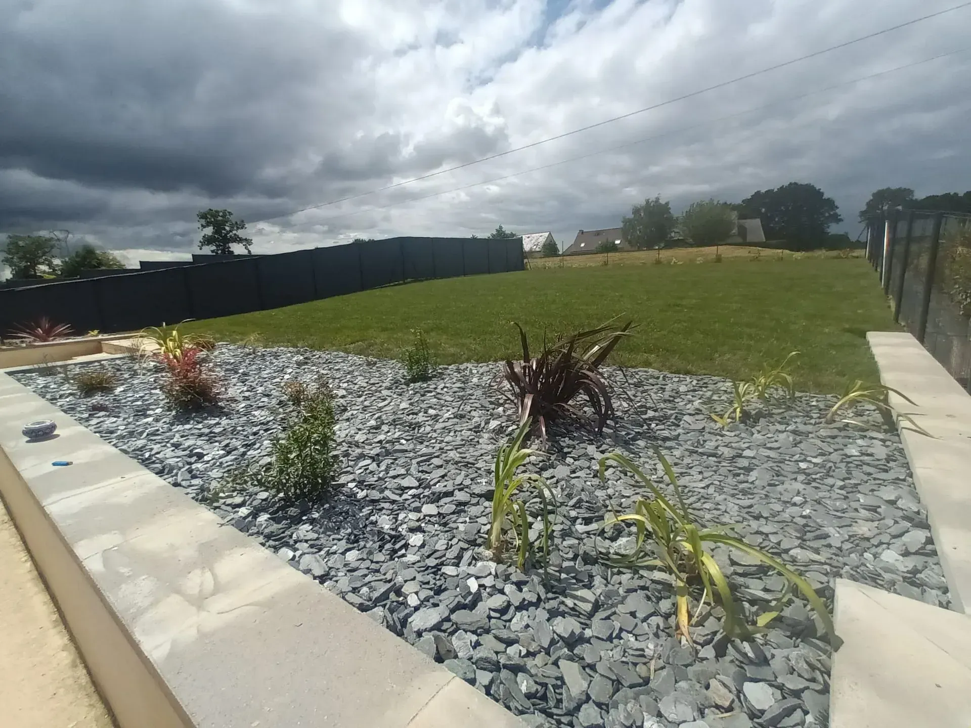 Un jardin paysager avec des rochers gris, des plantes et de l'herbe verte. Ciel couvert avec une clôture sombre en arrière-plan.