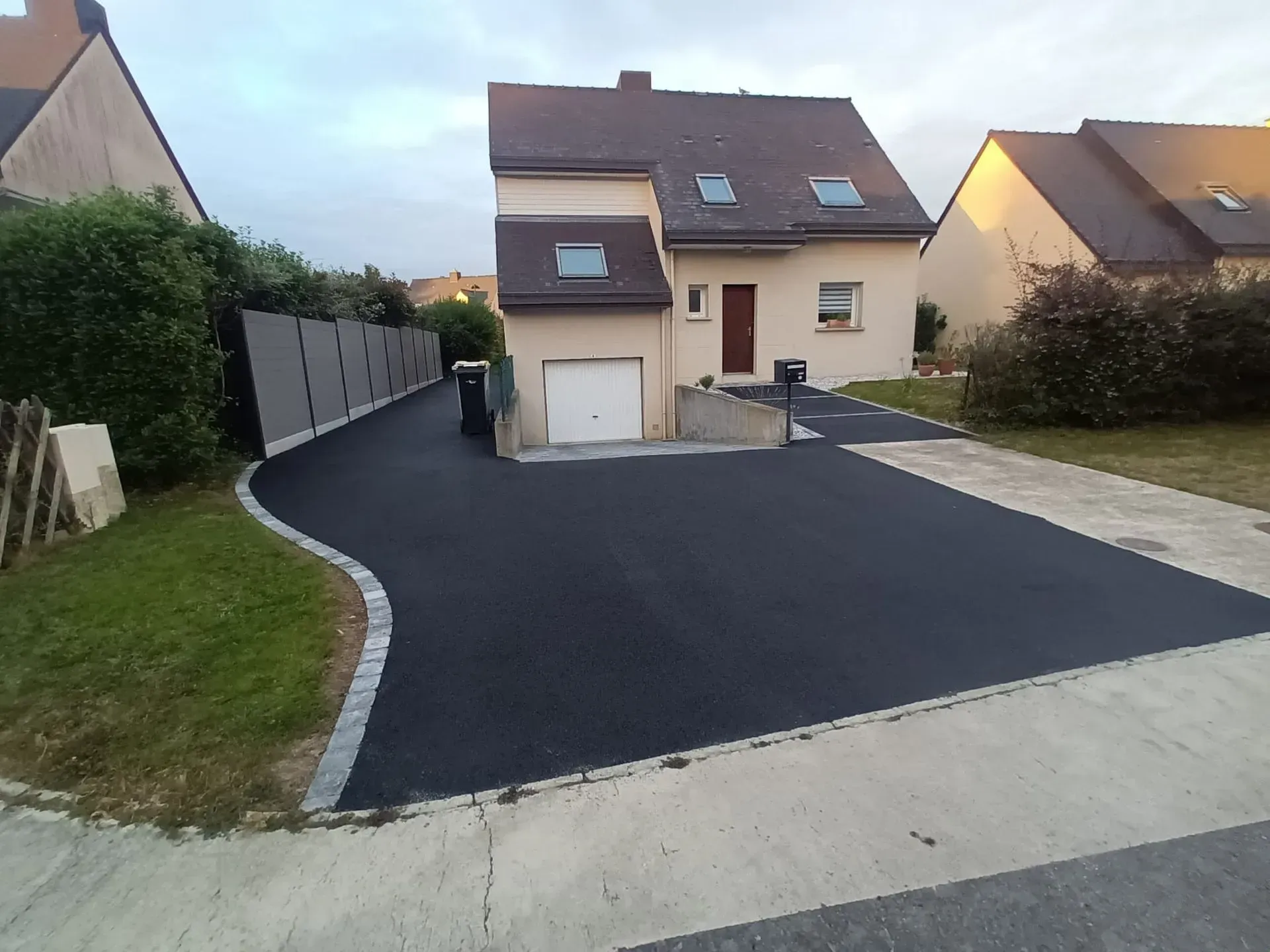 Allée asphaltée devant une maison beige à deux étages avec garage, bordée d'herbe verte et d'arbustes.