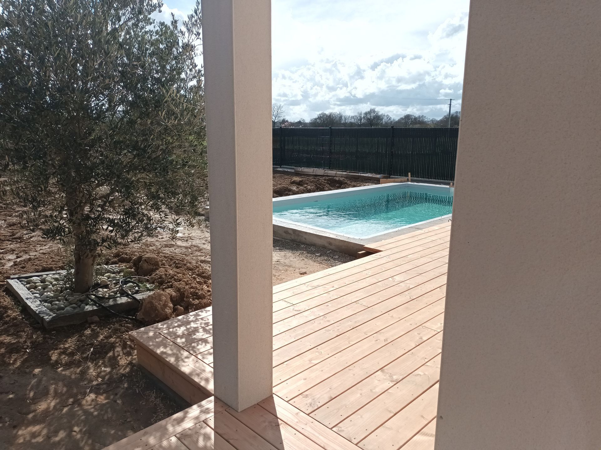 Olivier, terrasse partiellement construite et piscine visibles entre des piliers blancs par une journée ensoleillée.