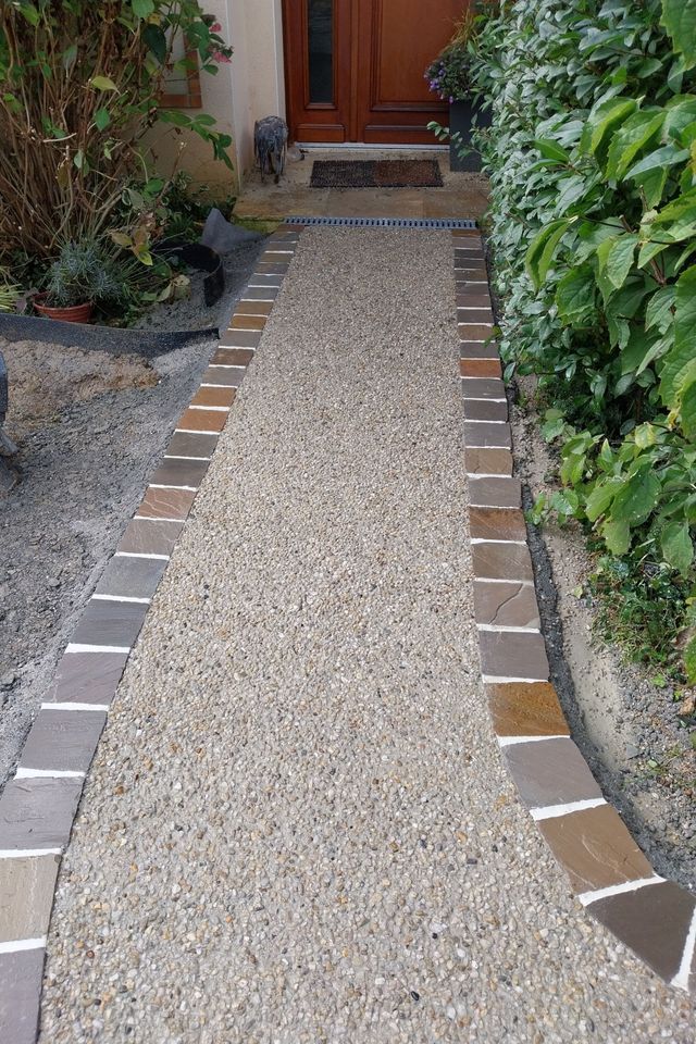 Un chemin de gravier bordé de tuiles marron et beiges mène à une porte en bois.