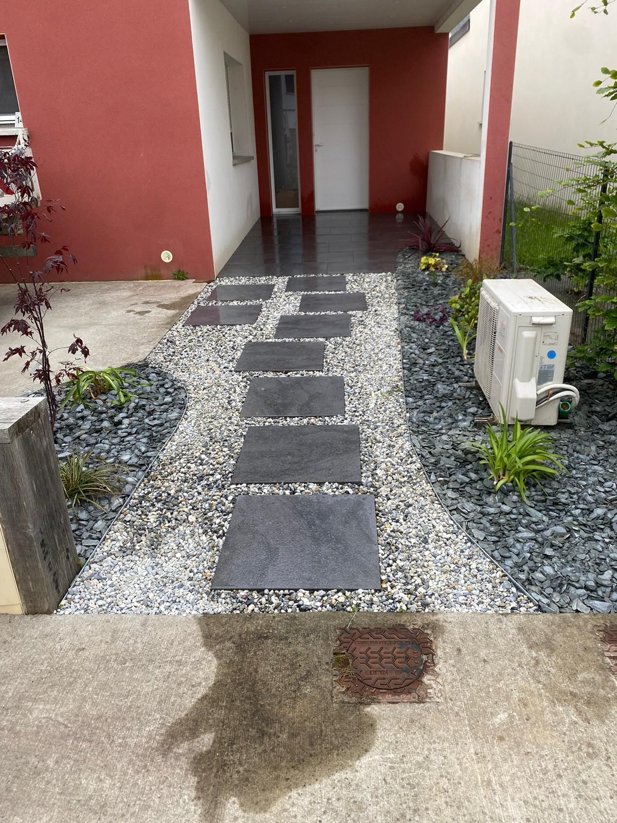 Des dalles de pierre sombres mènent à l'entrée d'un bâtiment rouge, flanquée de gravier et de plantes.
