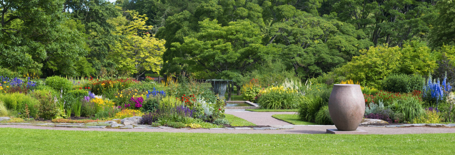 Jardin fleuri et coloré avec un chemin de pierre, une végétation dense et un grand pot en terre cuite au premier plan.