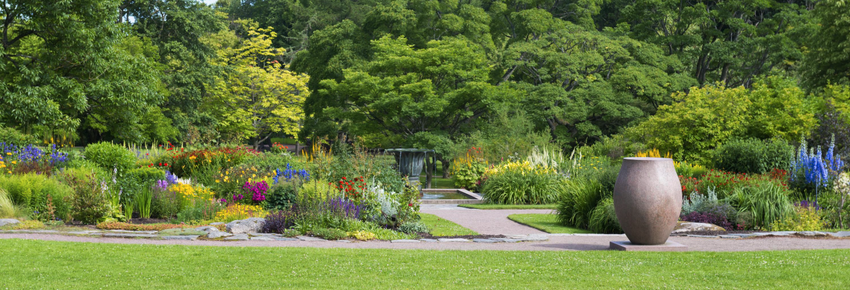 Jardin fleuri et coloré avec un chemin de pierre, une végétation dense et un grand pot en terre cuite au premier plan.