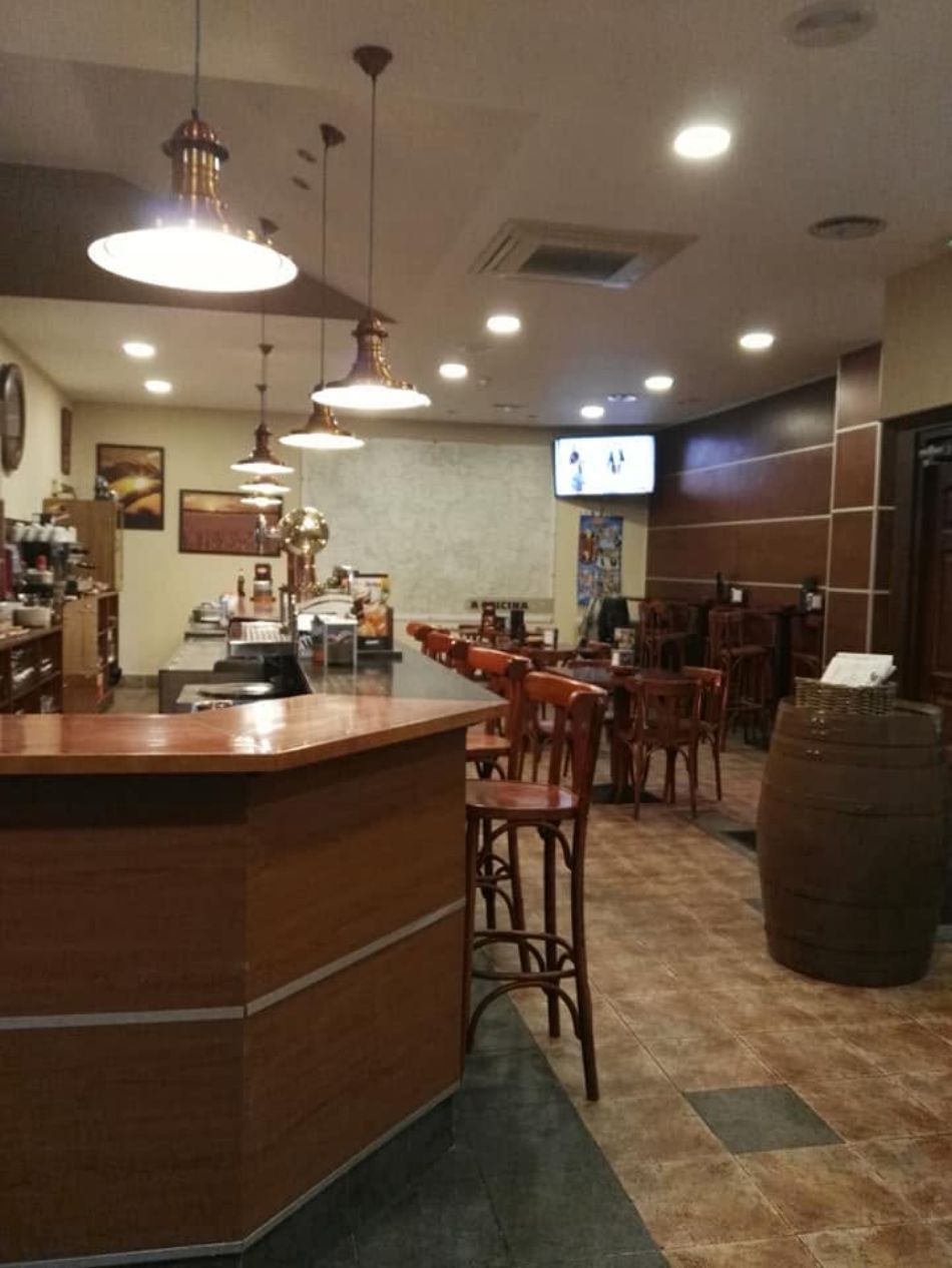 Interior acogedor de una cafetería con barra de madera, taburetes, lámparas colgantes y un televisor al fondo.