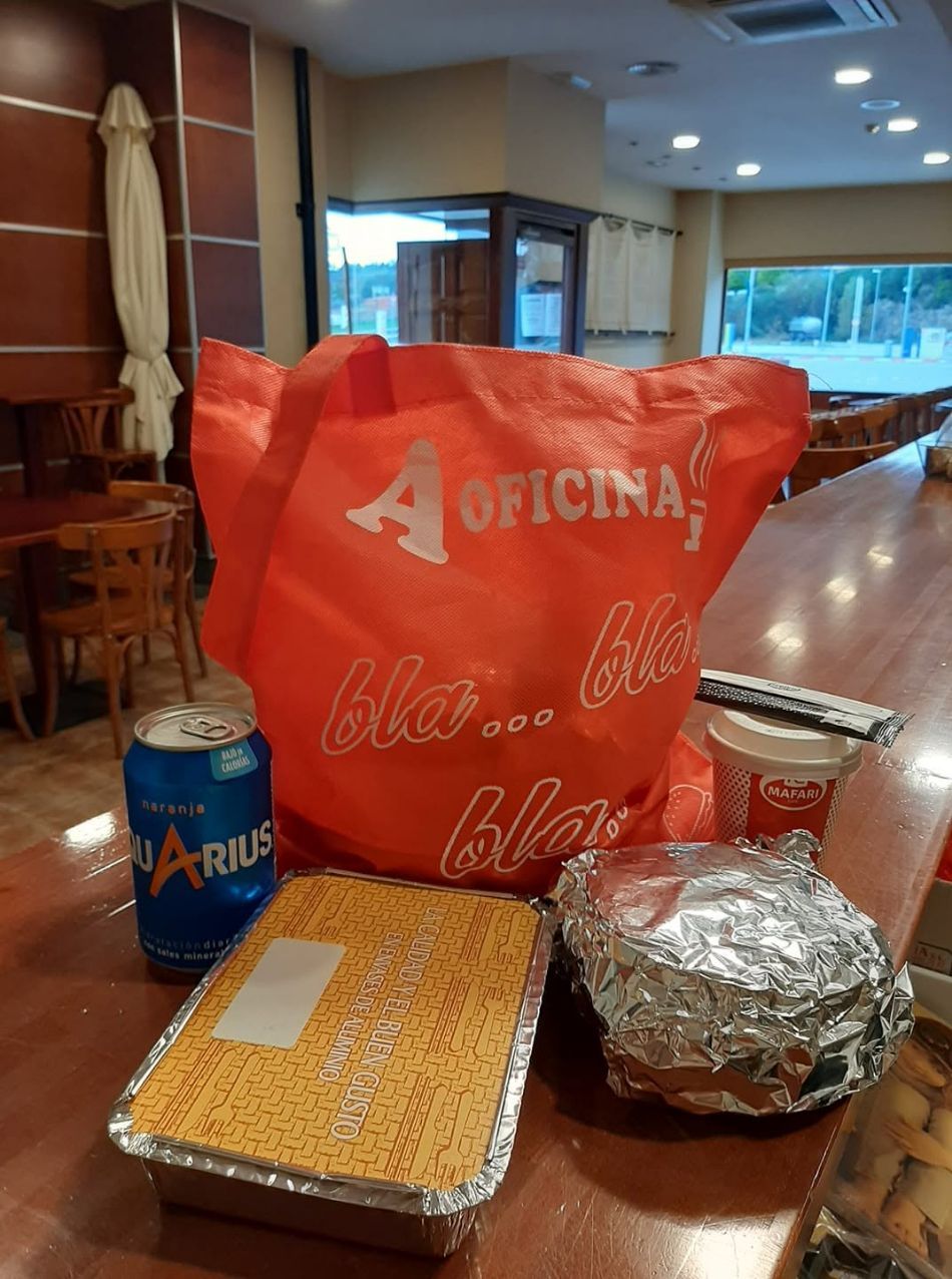 Mesa con una bolsa de la compra roja, bandejas de comida envueltas, una lata azul y un plato cubierto con papel de aluminio en un restaurante.
