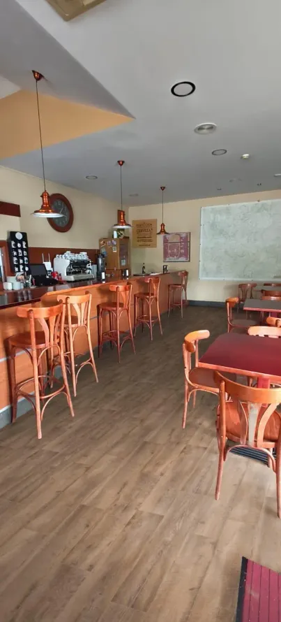 Interior vacío de una cafetería con barra de madera, taburetes, lámparas colgantes y un televisor al fondo.