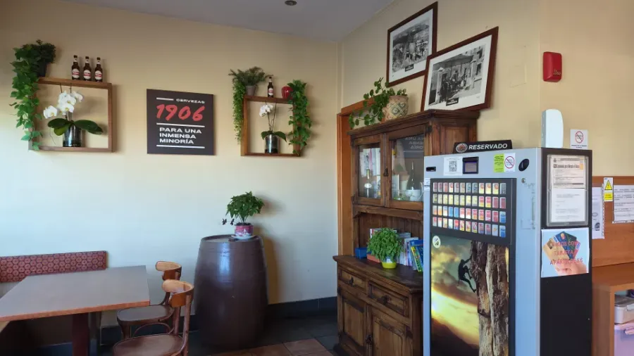 Zona de espera del restaurante con banco, armario de madera, plantas de pared, cuadros y máquina expendedora.