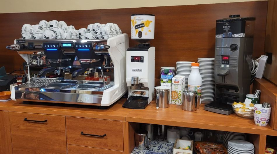 Estación de café con máquina de espresso, molinillo, tazas, jarabes y accesorios sobre un mostrador de madera.