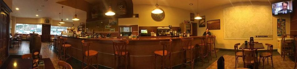 Interior de bar con iluminación cálida, barra de madera, lámparas colgantes, taburetes, mesas y un pequeño televisor en la pared.