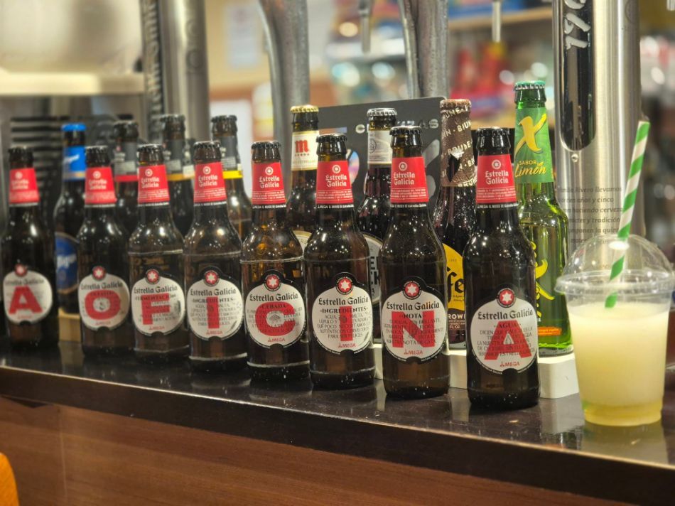 Botellas de cerveza que forman la palabra “AOFICINA” en la barra de un bar, con una bebida de color amarillo pálido al lado.