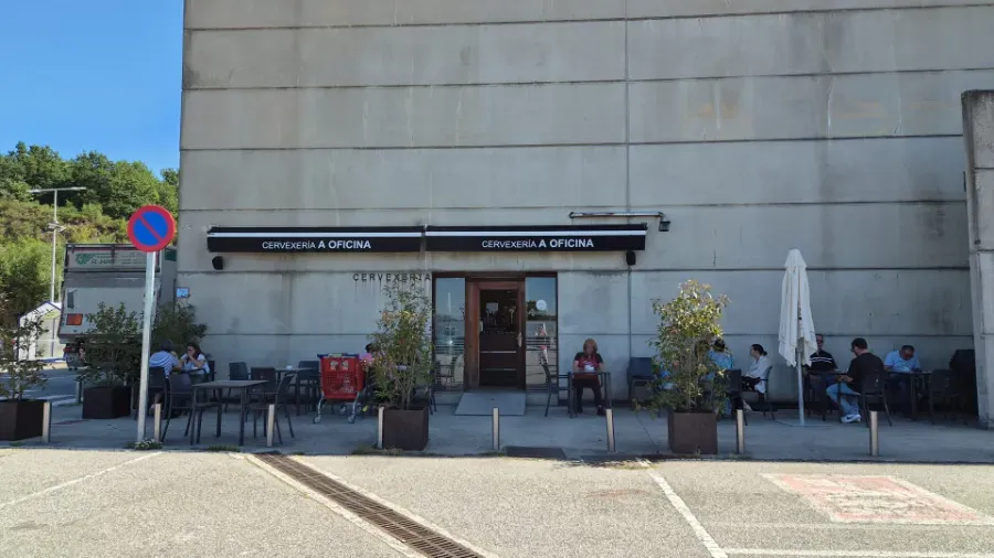 Cafetería al aire libre con mesas y sillas, situada fuera de un edificio, bajo un letrero negro y una gran sombrilla blanca.