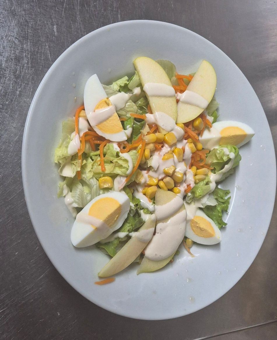 Ensalada en un plato blanco con lechuga, manzanas en rodajas, gajos de huevo, maíz y aderezo sobre una encimera de acero inoxidable.