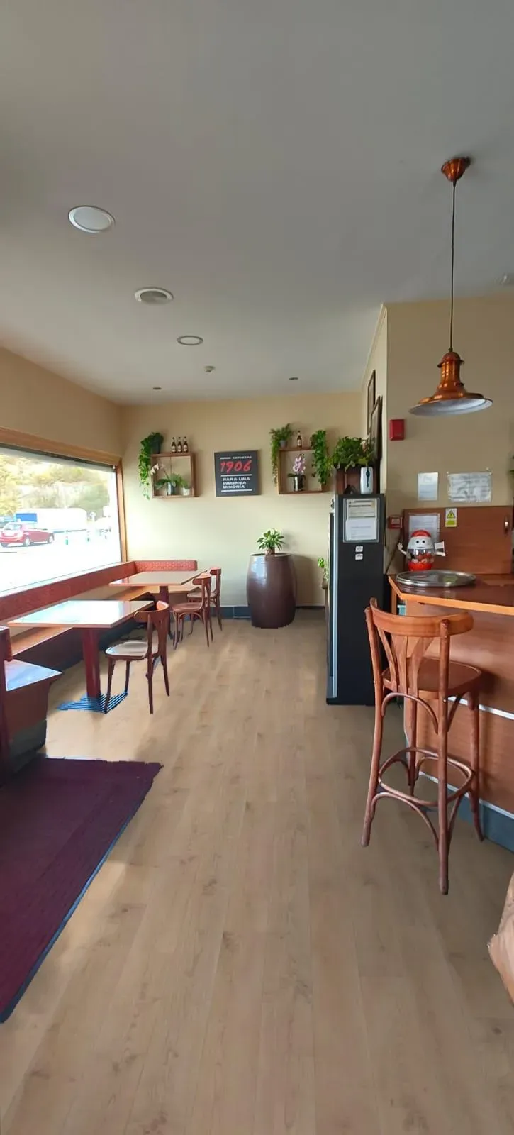 Interior luminoso de una cafetería con mesas y sillas de madera, lámparas colgantes y plantas en macetas a lo largo de un pasillo.