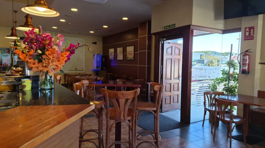 Interior acogedor de una cafetería con mesas de madera, flores coloridas y una puerta abierta con vistas a una calle luminosa.