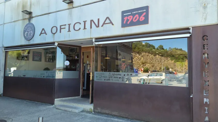Fachada de “A Oficina 1906” con ventanas de cristal y puerta abierta en una calle soleada.