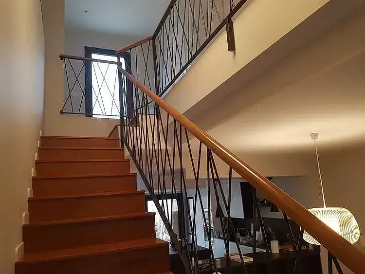 Escalera interior con escalones de madera, barandilla de hierro y una lámpara en un luminoso interior de una casa.