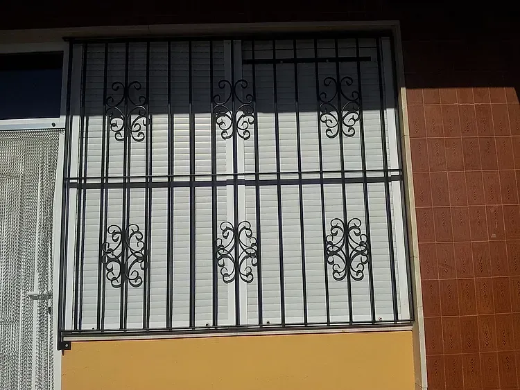 Rejas de hierro forjado negro sobre contraventanas blancas en la fachada de un edificio de color beige.