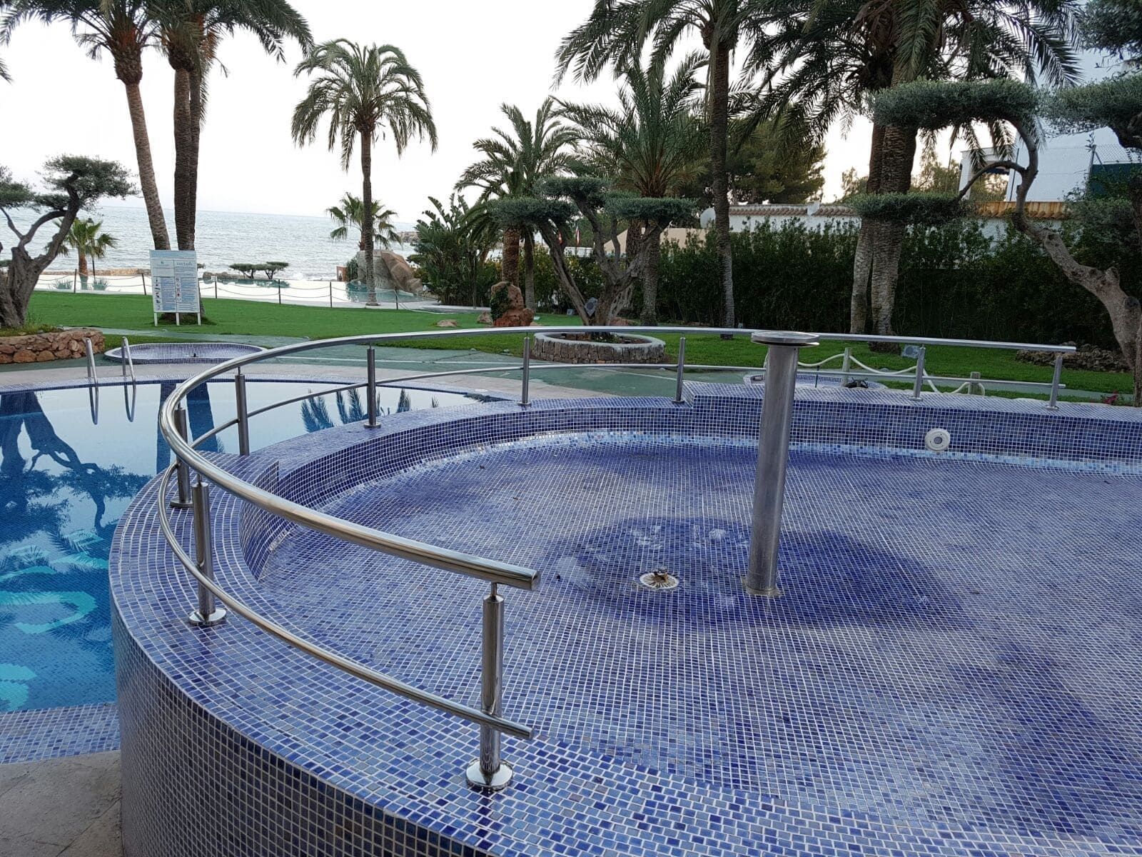 Spa circular con azulejos y pasamanos de metal junto a una piscina, con vistas a las palmeras y al océano.