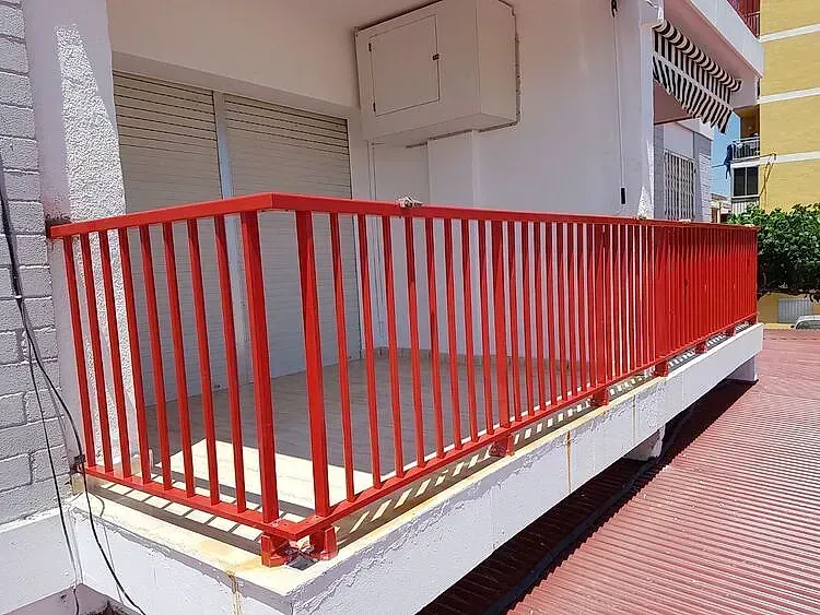 Barandilla de metal rojo alrededor de un pequeño balcón con paredes blancas y un pasillo de baldosas rojas al lado.