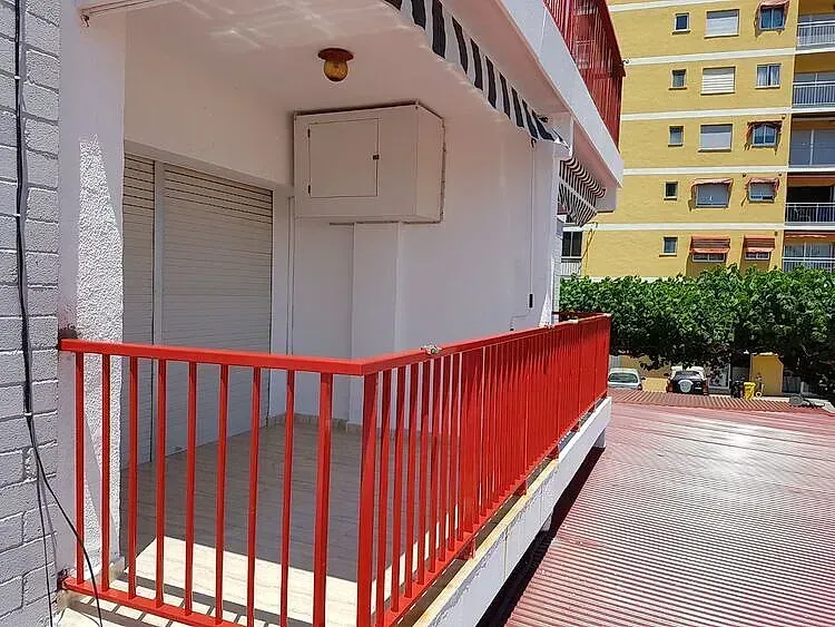 Pasarela con barandilla roja junto a un edificio con contraventanas cerradas y un patio con baldosas debajo.