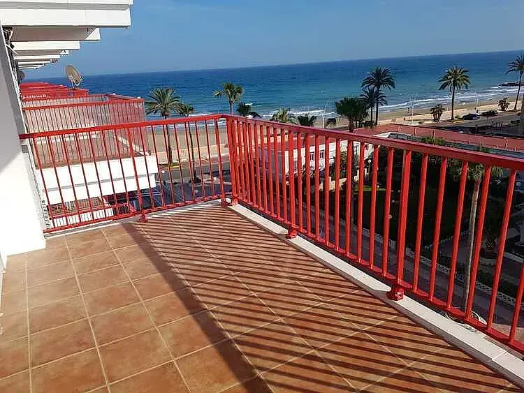 Balcón con barandilla roja con vistas a una playa soleada y al océano.