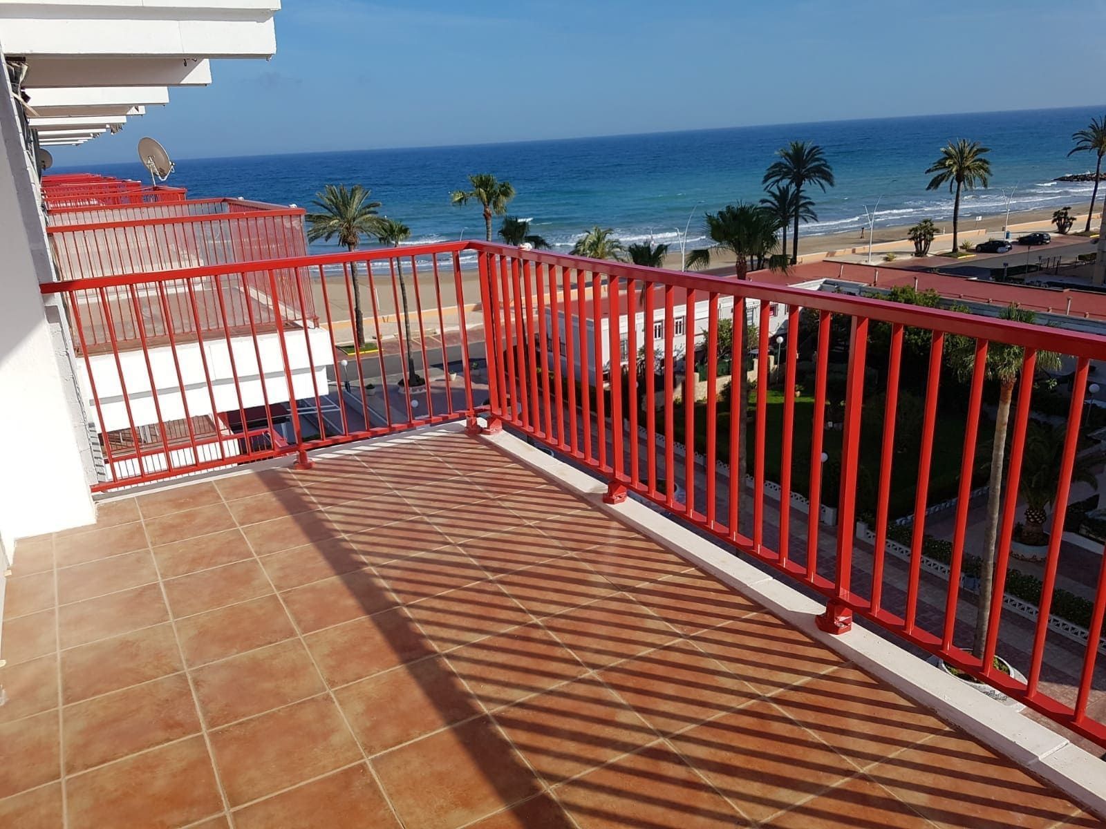 Terraza junto al mar con barandilla roja, suelo de baldosas y vistas al océano bajo un cielo azul despejado.