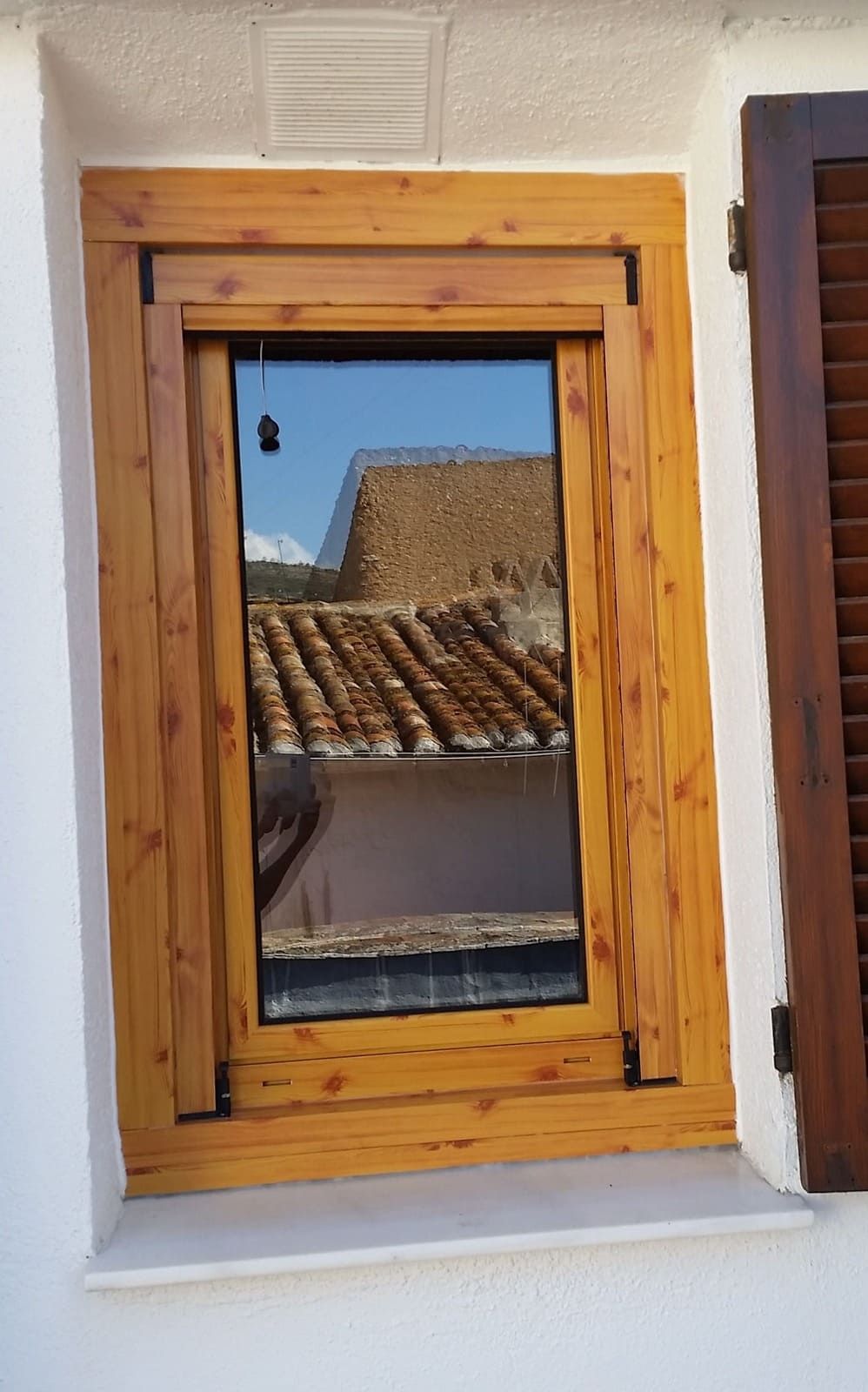 Ventana con marco de madera que refleja un tejado de tejas y el cielo azul en una pared blanca.