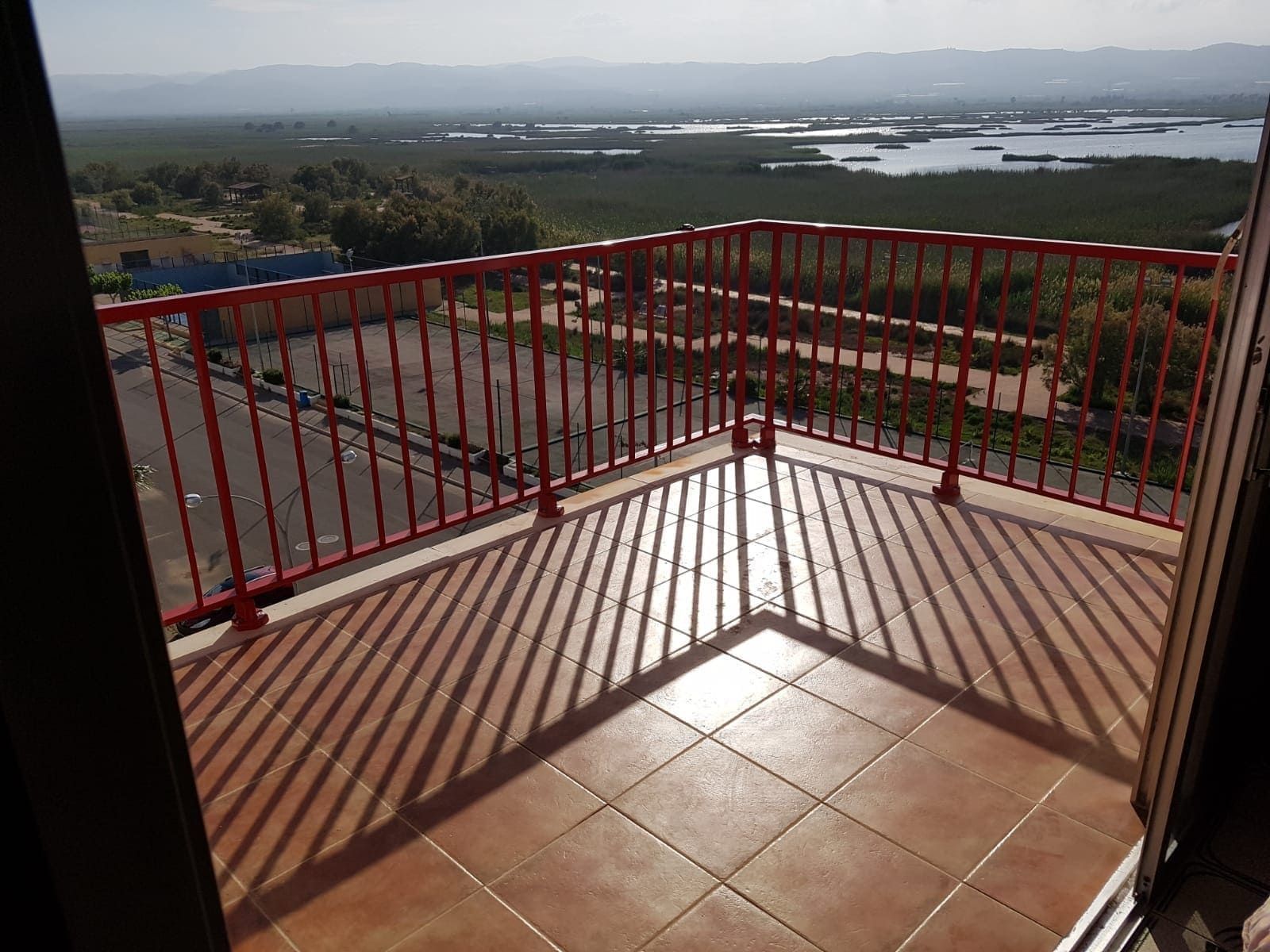 Balcón con barandillas rojas y suelo de baldosas con vistas a un paisaje soleado y al agua a lo lejos.