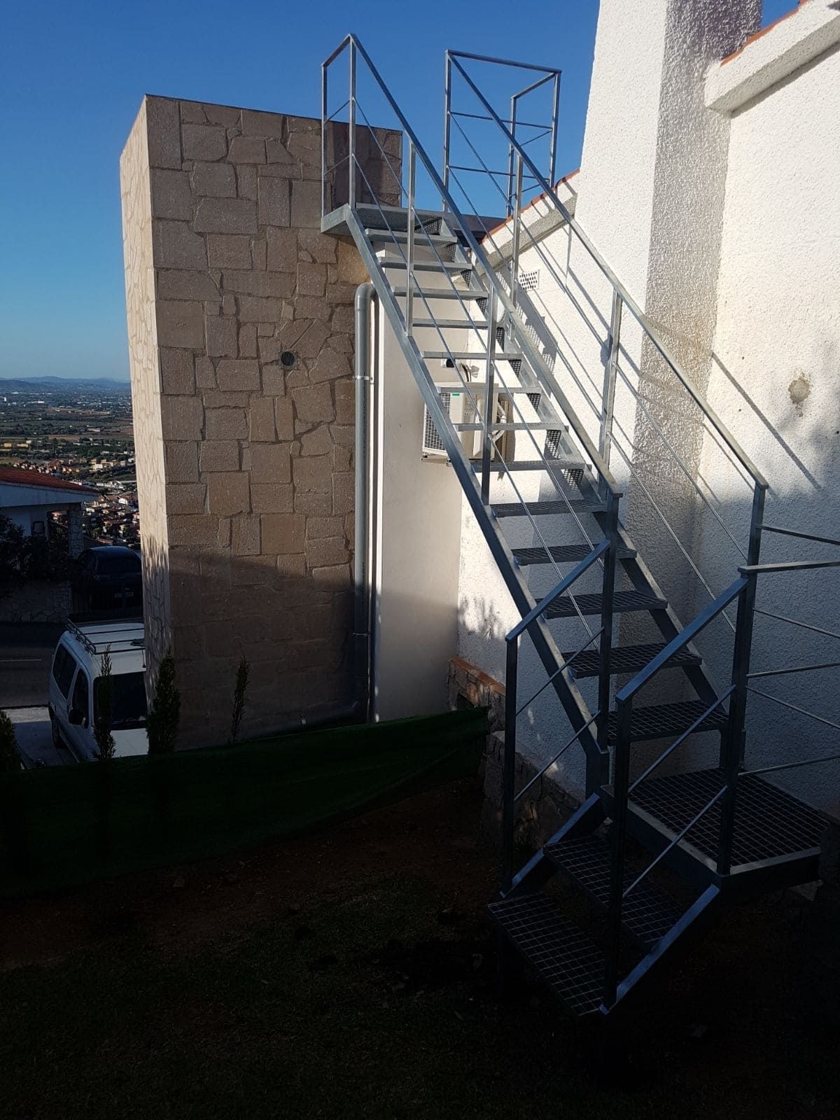 Escalera metálica exterior junto a un edificio blanco, con un muro de piedra y vistas a la ciudad al fondo.