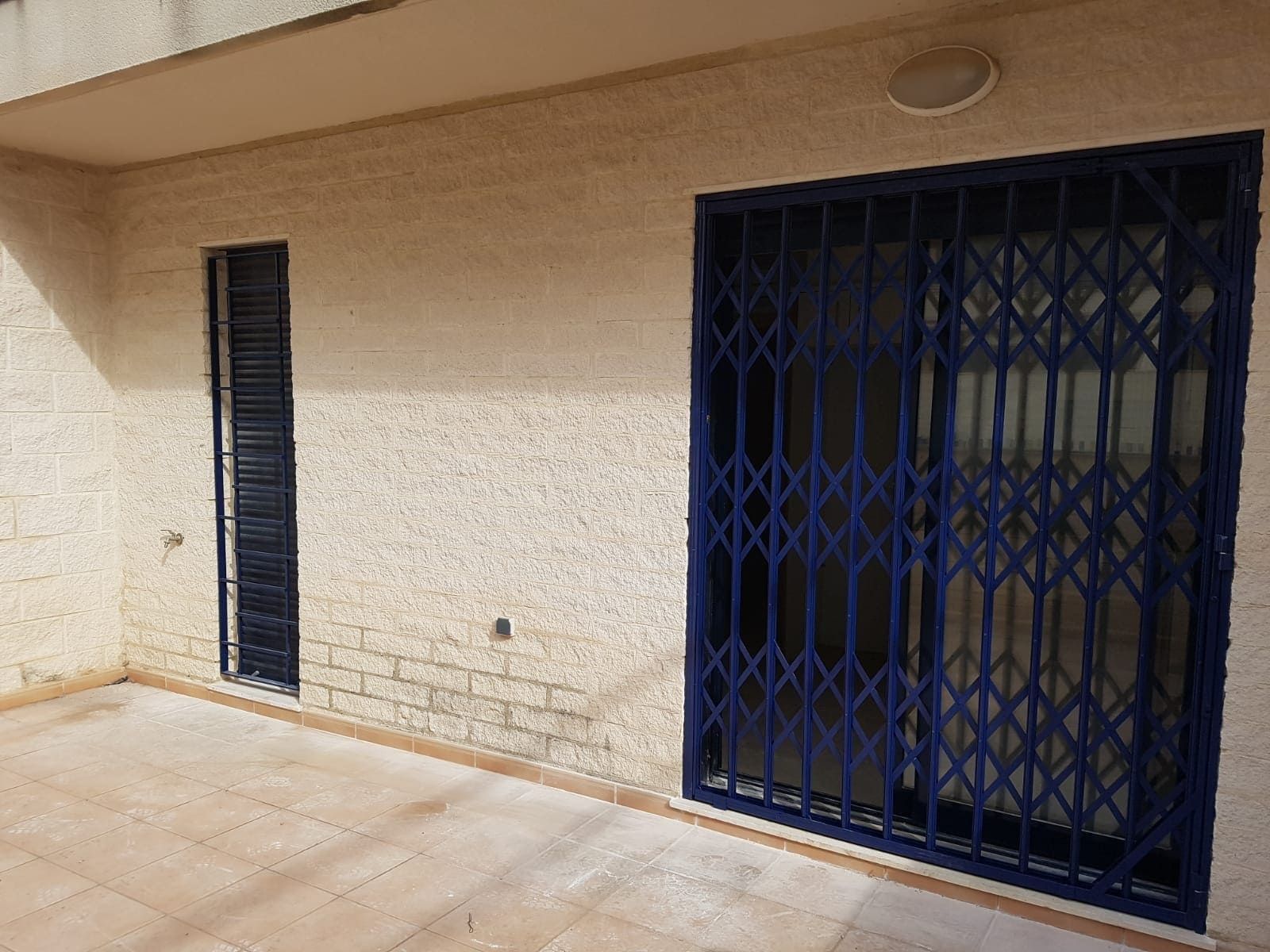 Pared exterior de color beige con una pequeña ventana negra y una puerta de seguridad azul en un patio embaldosado.