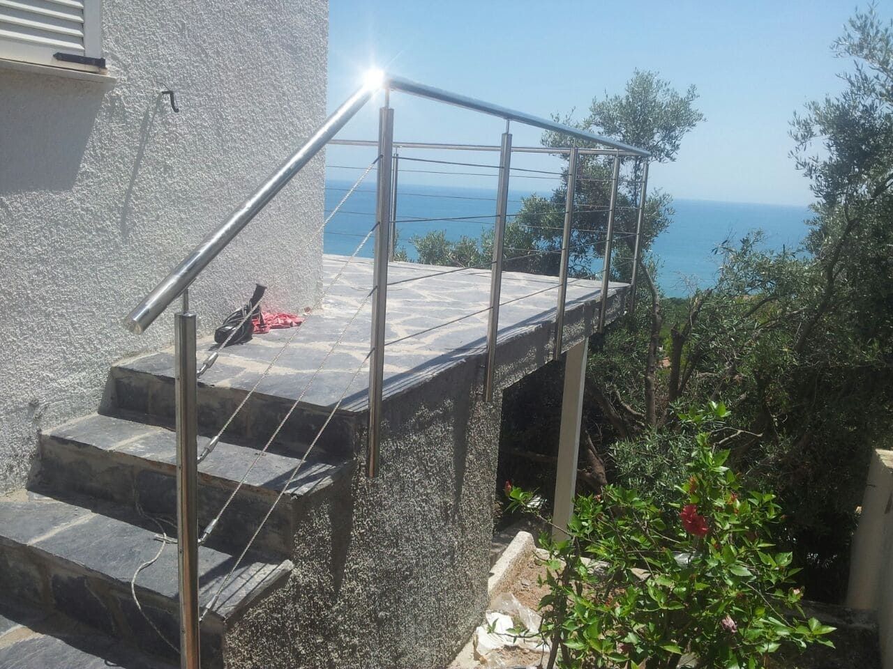 Escaleras de hormigón que conducen a una terraza junto al mar con barandilla metálica, con vistas al océano azul y a la vegetación.